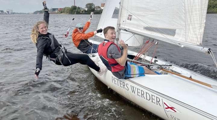 Drei Personen segeln lachend auf einer Jolle des Segelvereins Weser in Bremen. Eine Frau hängt im Trapez seitlich über dem Wasser, während die beiden anderen Crewmitglieder im Boot sitzen und in die Kamera winken. Das Boot trägt den Namen ‚Peters Werder‘ und fährt bei lebhaftem Wind über die Weser.
